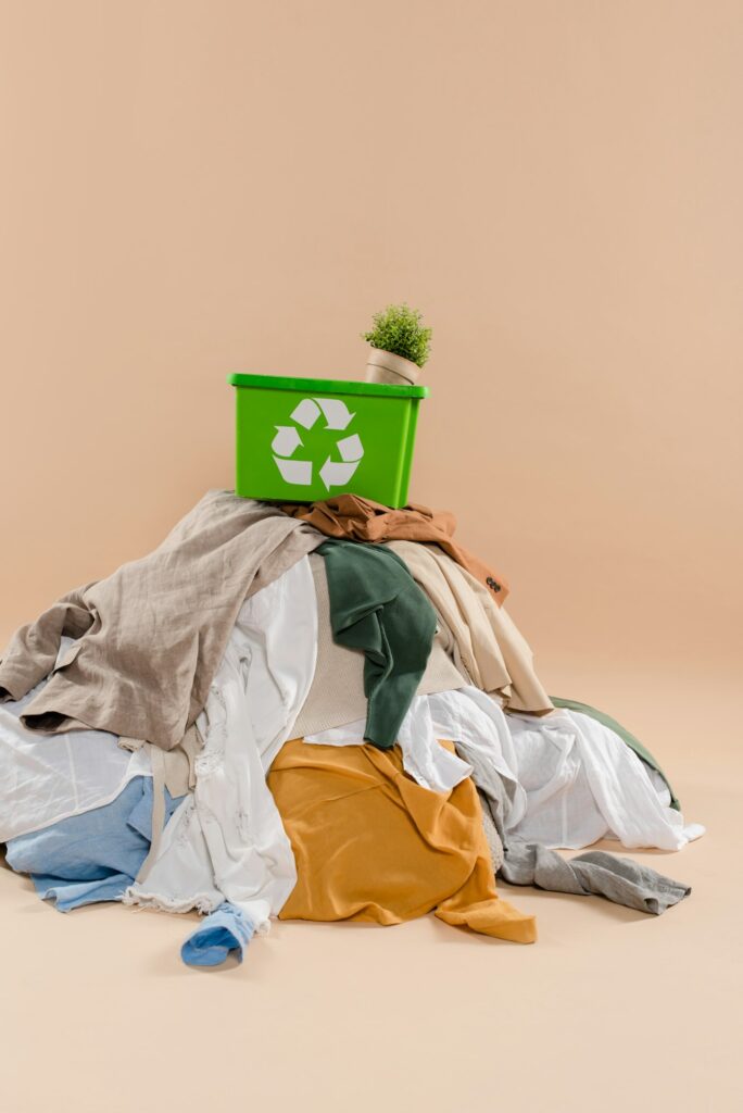 green recycling box with plant in pot on stack of clothing on beige background, environmental saving